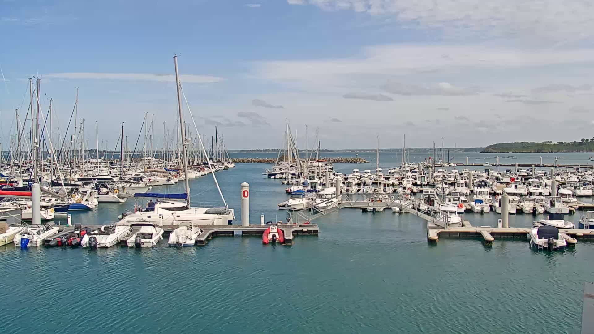A large marina filled with numerous sailboats and motorboats is shown under a mostly sunny sky with some clouds.