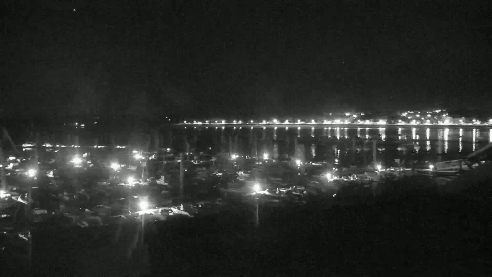 A nighttime view of a harbor filled with boats, with a town's lights reflecting on calm water in the background.
