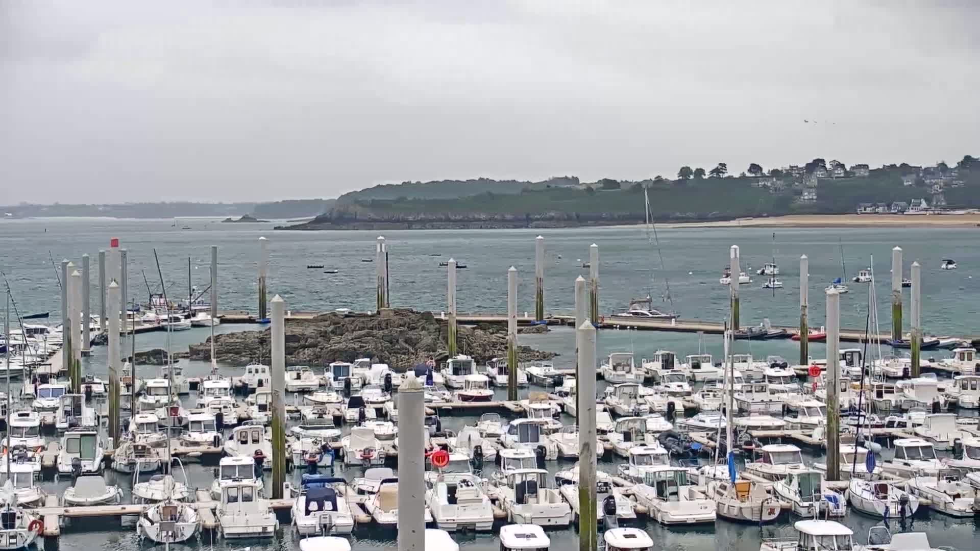 A marina filled with numerous boats is situated on a rocky outcrop, under an overcast sky, with a calm body of water and distant land visible in the background.