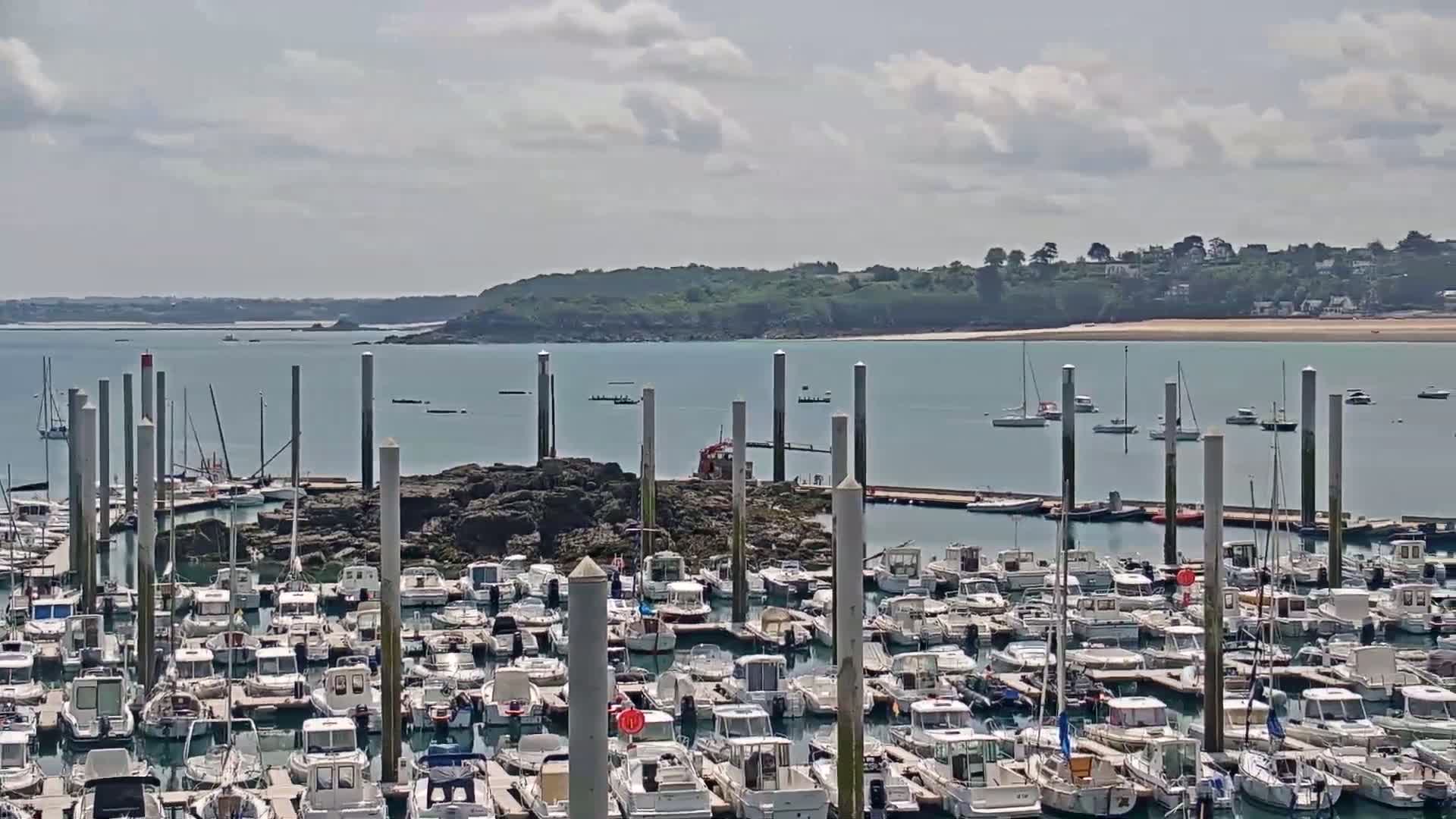 A marina filled with numerous boats is situated on a calm body of water under a partly cloudy sky, with a landmass visible in the distance.