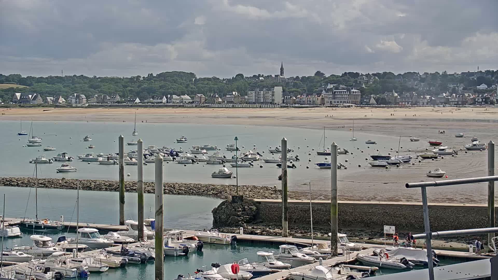 A coastal town with a sandy beach is visible across a harbor filled with many boats moored at docks under a partly cloudy sky.