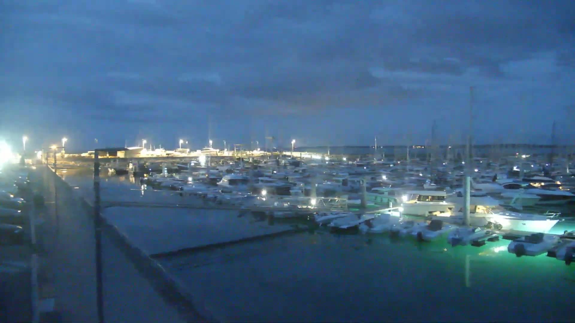A coastal town with a sandy beach is visible across a harbor filled with many boats moored at docks under a partly cloudy sky.