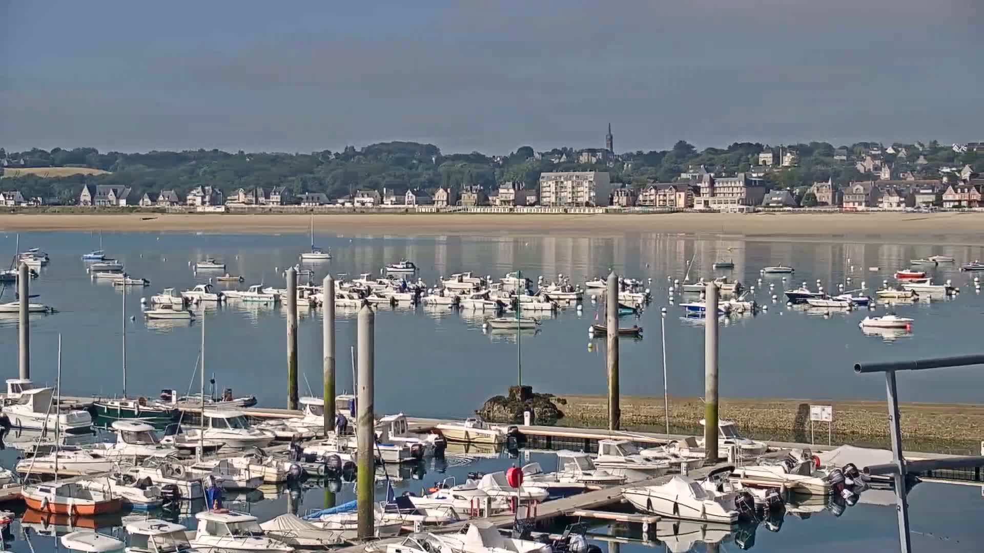 A calm bay filled with numerous boats at a marina, with a town and sandy beach visible in the background under a clear sky.