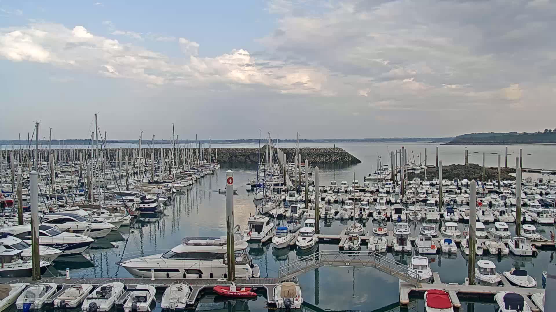 A large marina filled with many sailboats and motorboats is situated on calm water under a partly cloudy sky.