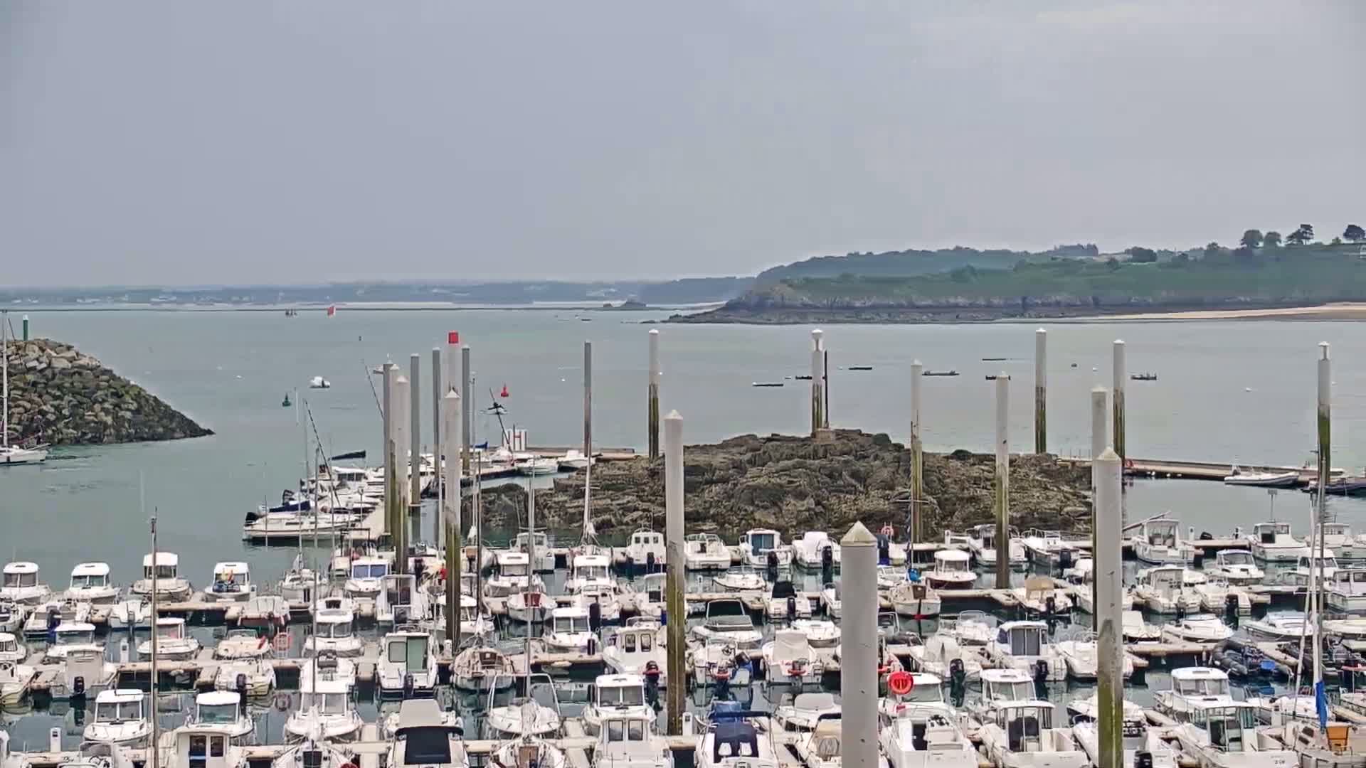 A harbor filled with numerous docked boats on a calm, sunny day, with a rocky outcrop in the center and a distant shoreline visible across the water.