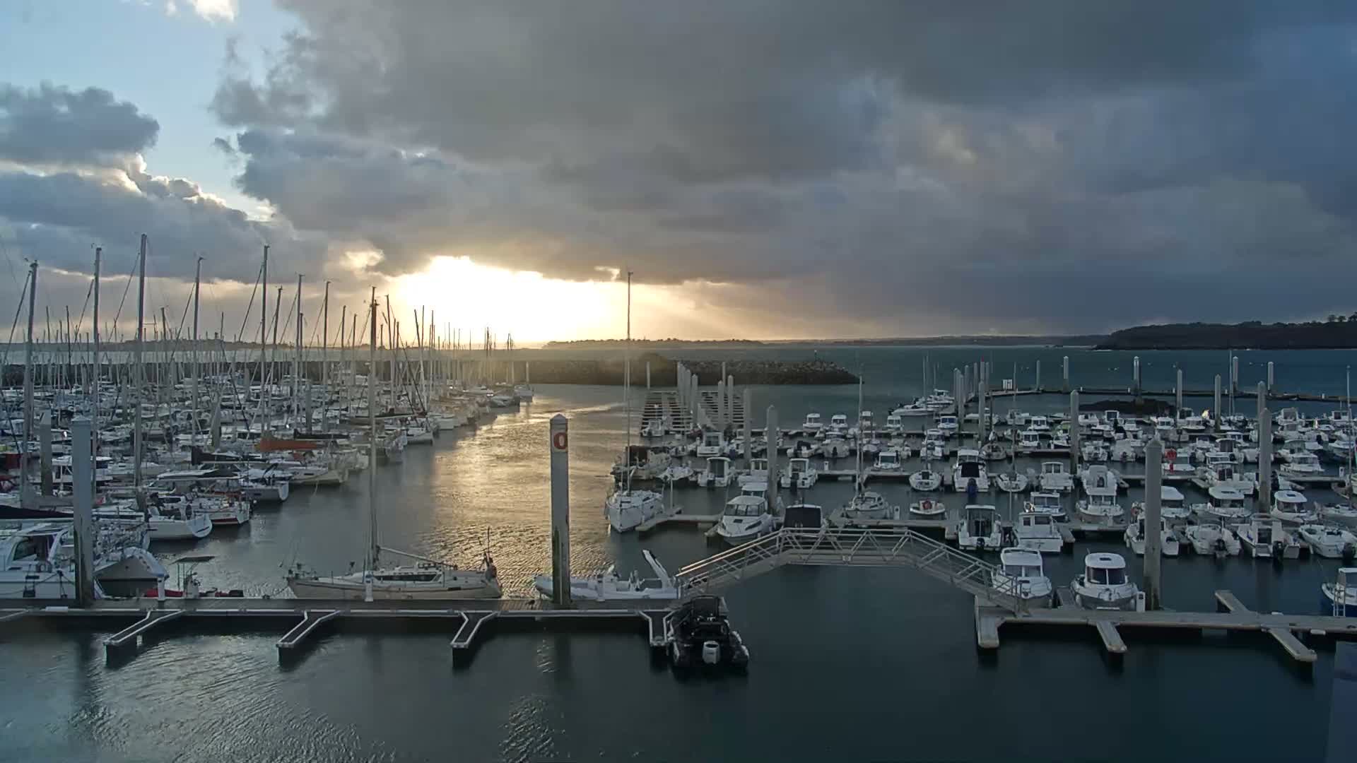 Numerous boats are docked in a large marina under a dramatic, heavily clouded sky, with a strong burst of golden sunlight piercing through the clouds and reflecting on the water.