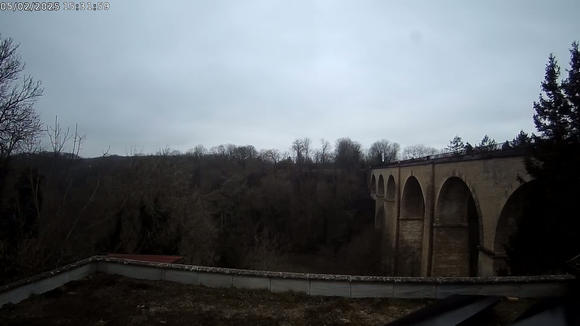 A stone viaduct with multiple arches is seen extending to the right, beyond a wall and a line of bare winter trees under an overcast sky.