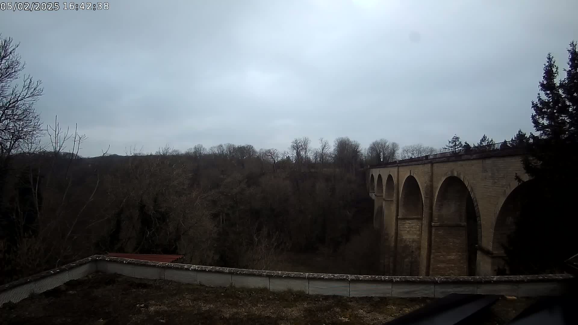 A stone bridge with multiple arches spans a wooded valley under an overcast sky.