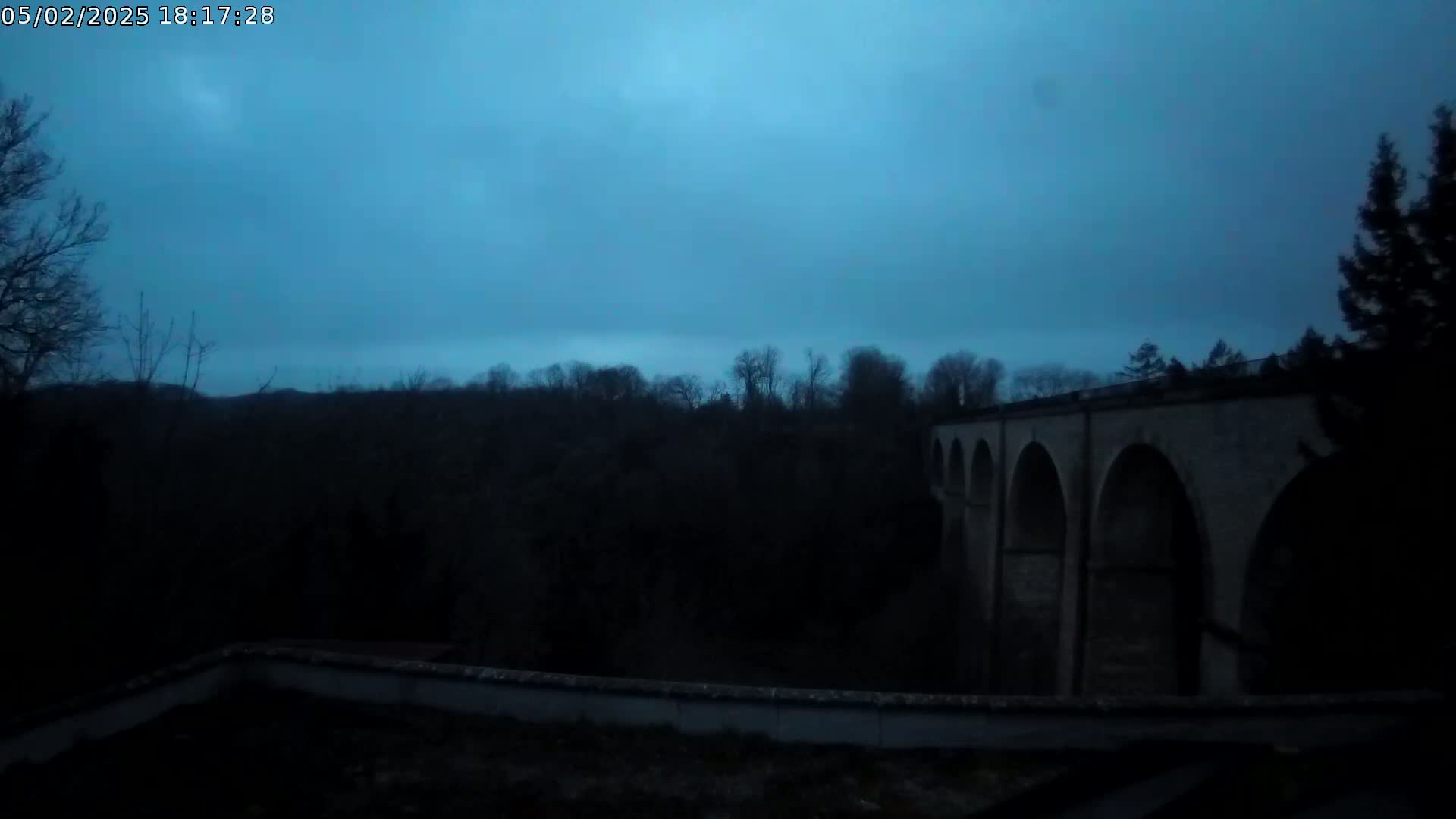 A large stone arched bridge is visible at dusk under a dark blue, overcast sky, set against a backdrop of silhouetted trees.