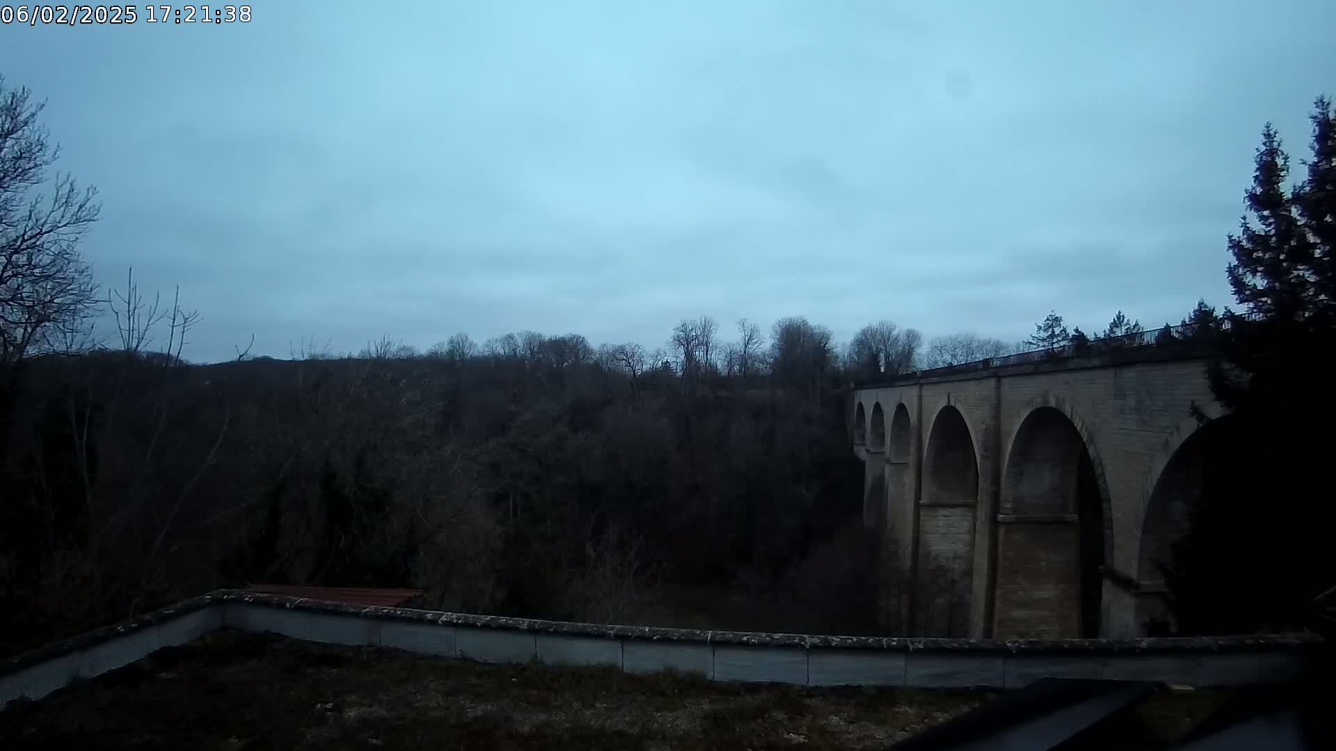 A stone viaduct with multiple arches is seen under an overcast sky, beyond a low wall and dark, leafless trees.