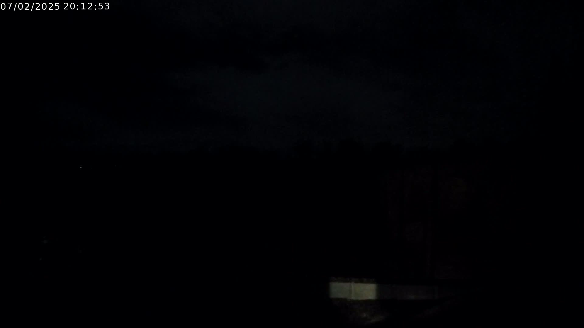 A dark, nighttime scene shows a dimly lit, low wall or barrier in the lower right corner under a mostly cloudy sky.