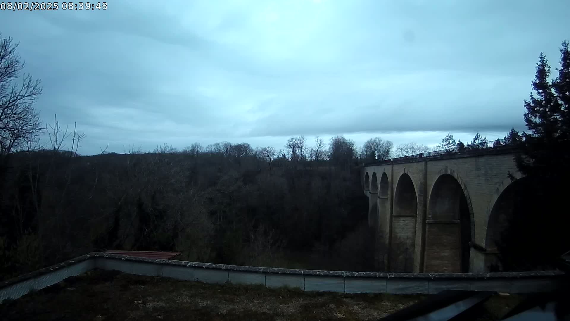 A stone bridge with multiple arches spans a wooded valley under a cloudy sky.