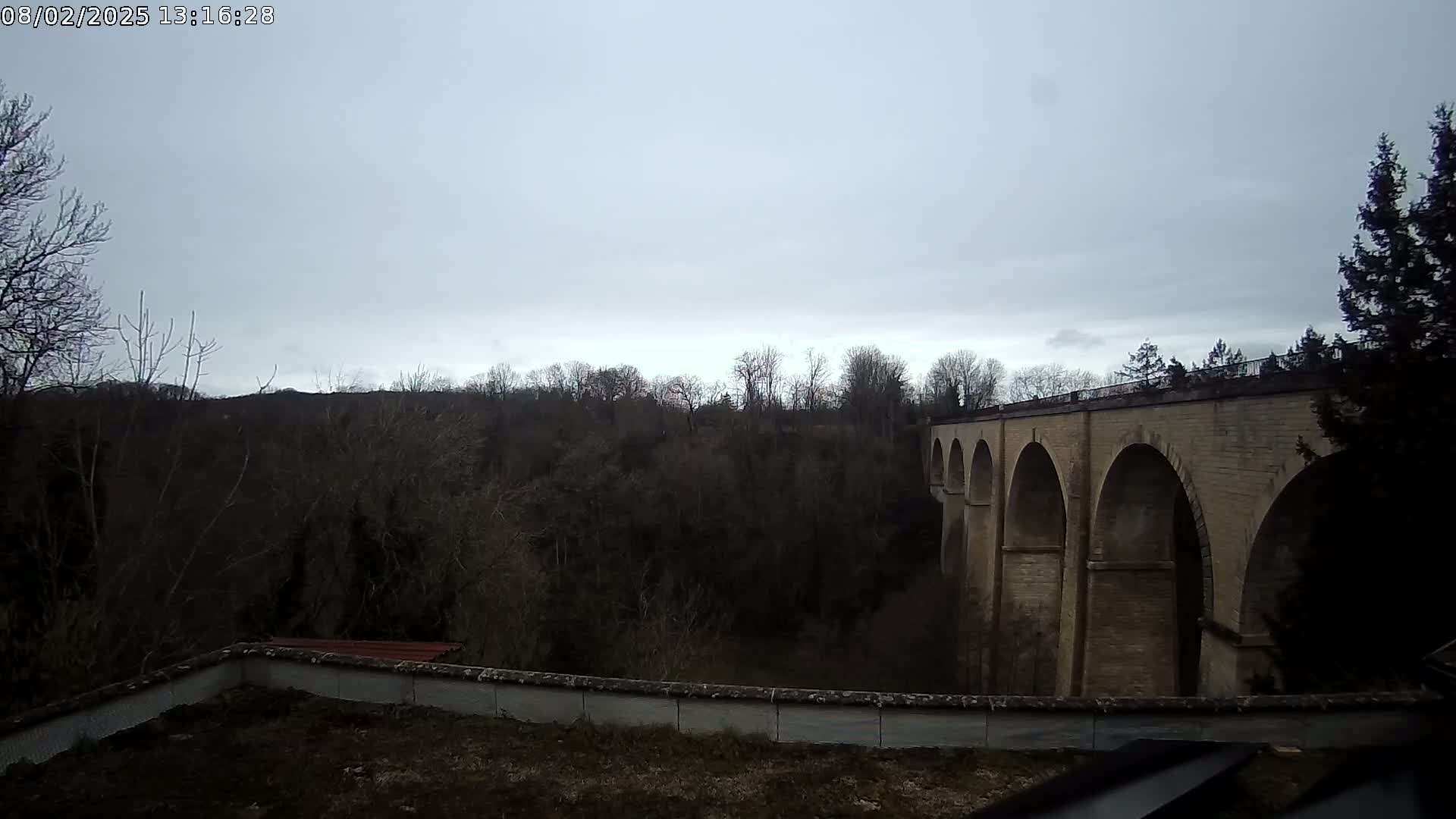 A stone viaduct with multiple arches is seen from a distance under an overcast sky, set against a backdrop of leafless trees.