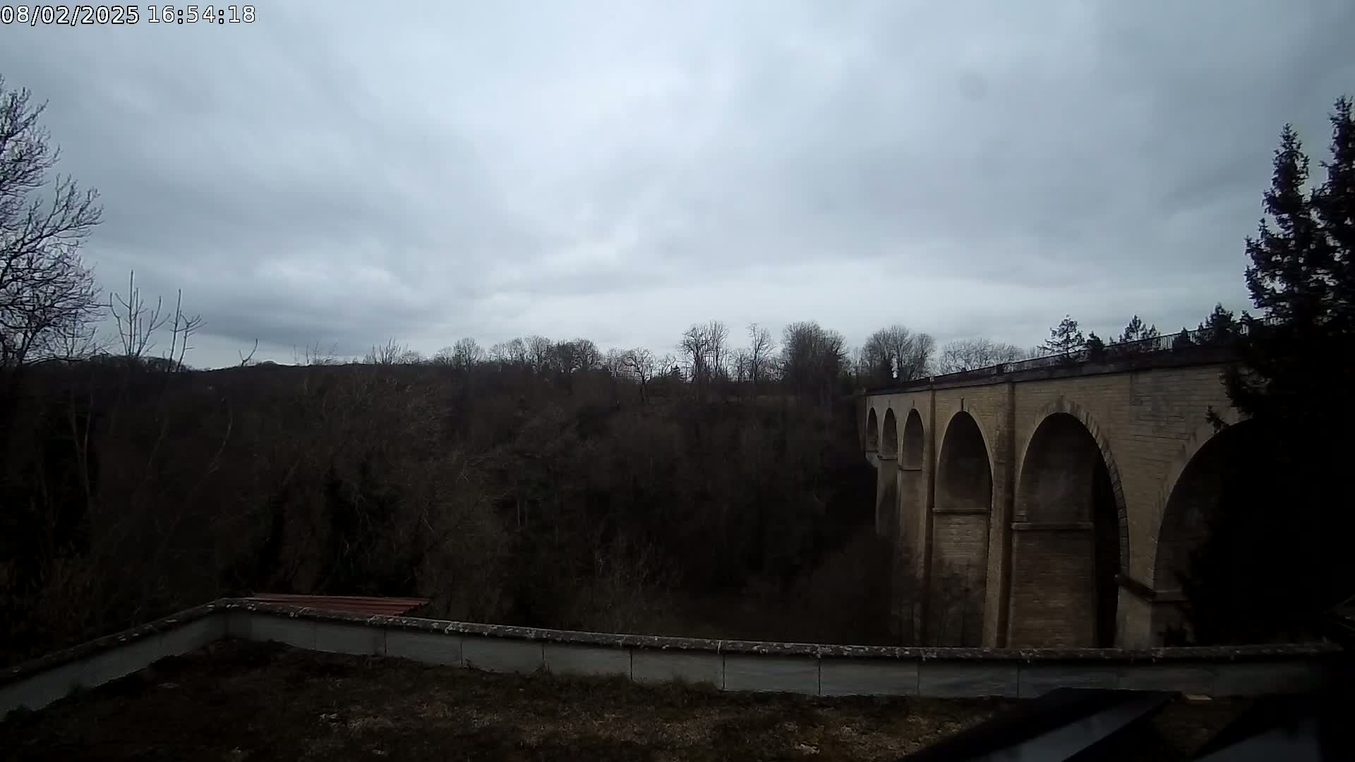 A stone arched bridge stretches across a wooded valley under a cloudy sky.