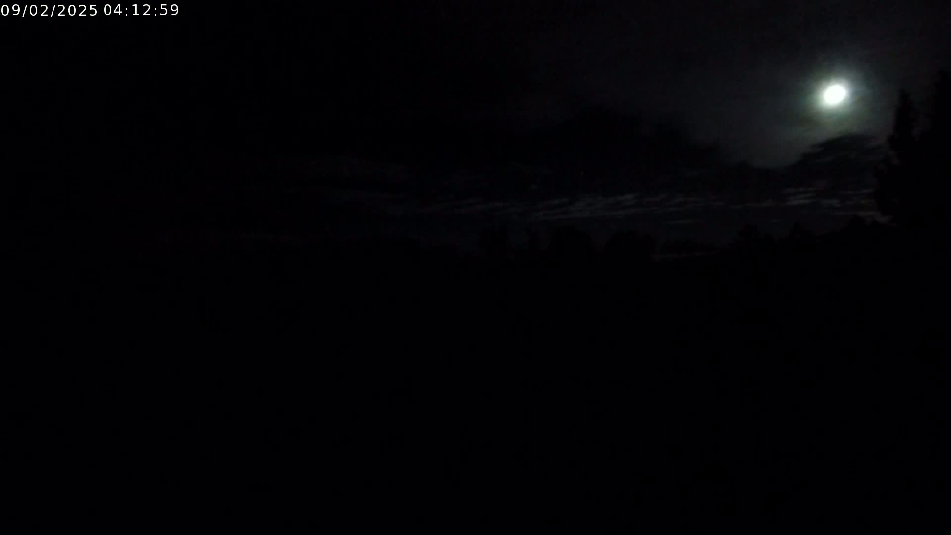 A bright moon shines through a mostly cloudy night sky above a dark, silhouetted landscape.