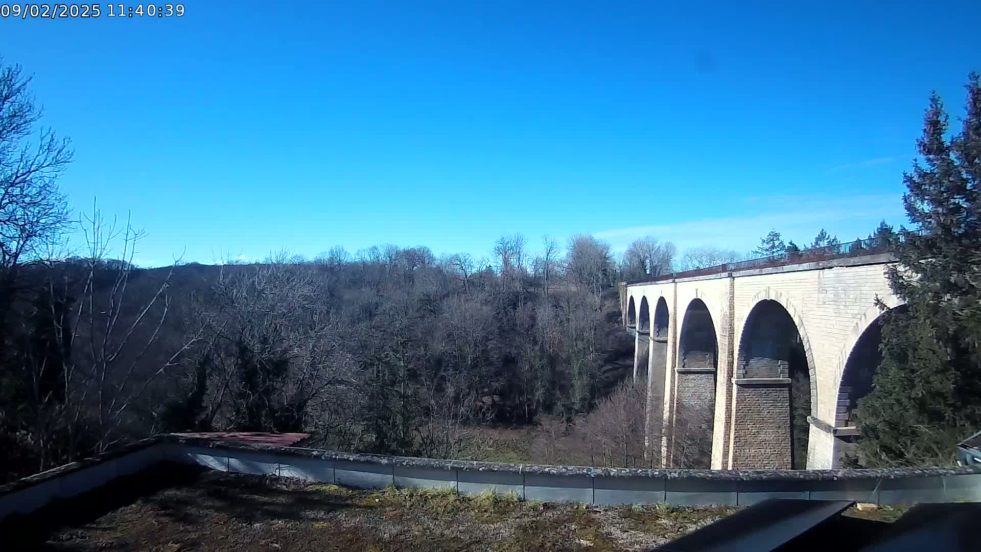A long stone viaduct with multiple arches stretches across a valley of leafless trees under a clear blue sky.