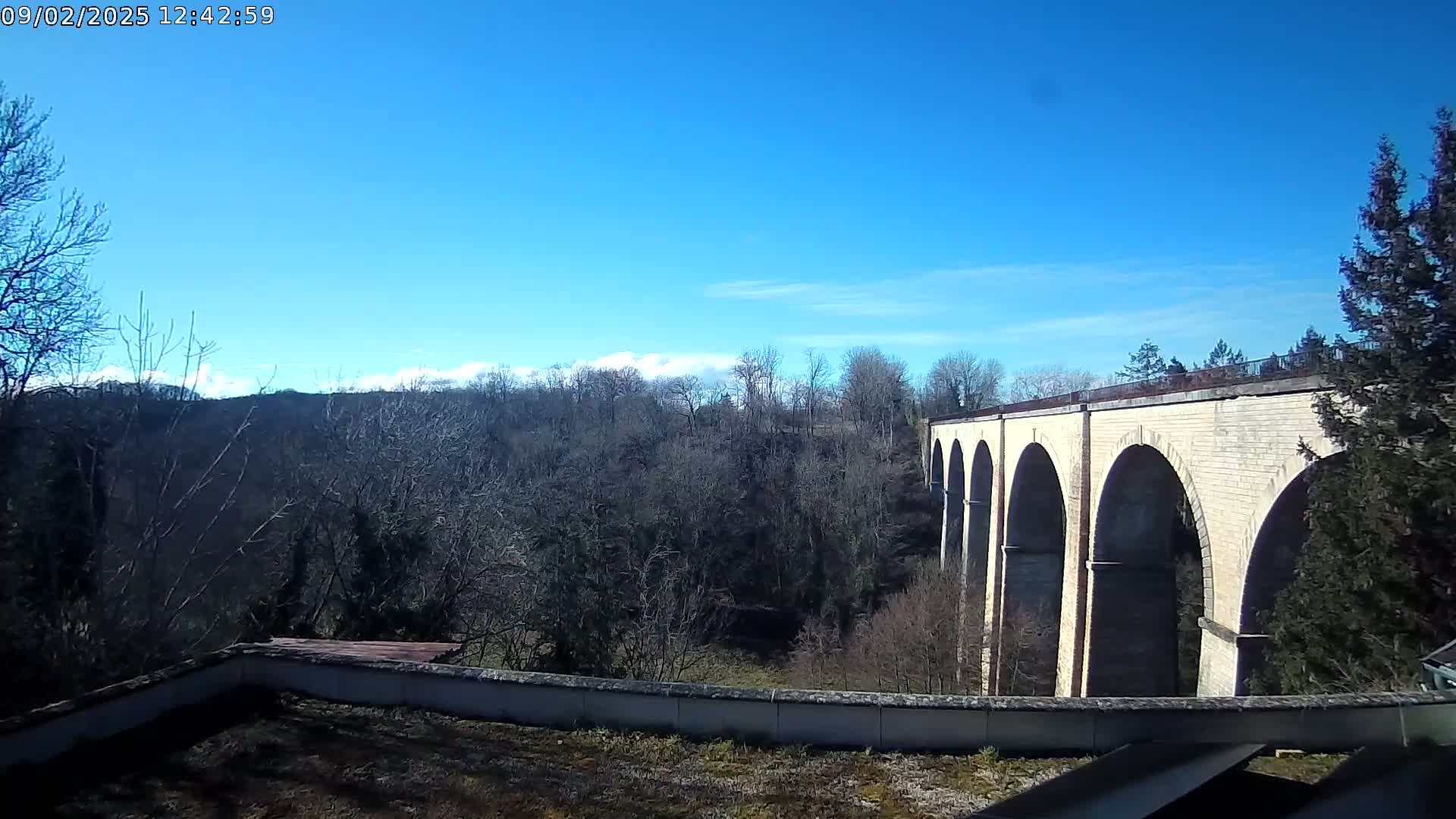 A stone viaduct with multiple arches stretches across a valley of leafless trees under a clear blue sky.