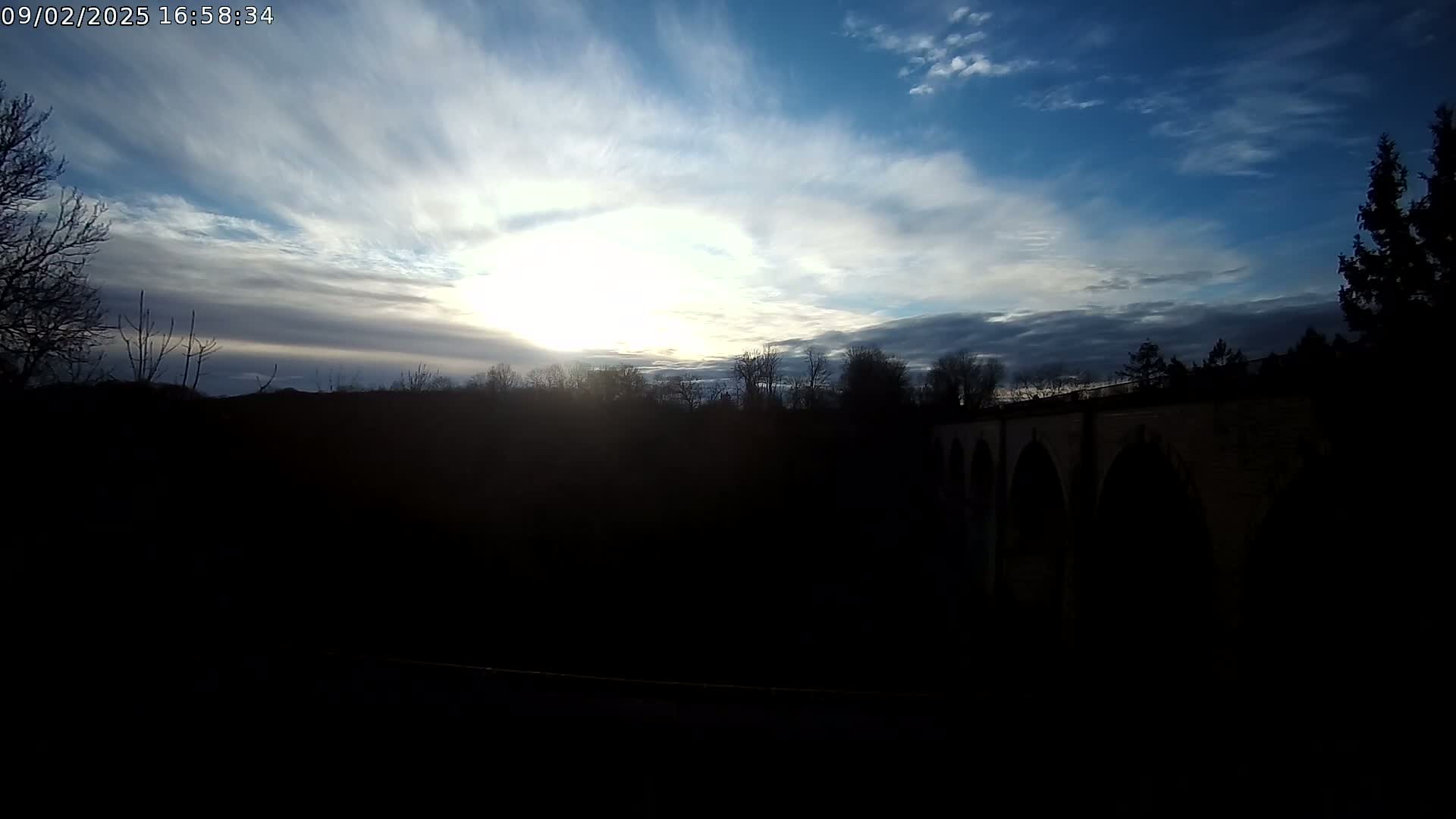 A stone bridge is silhouetted against a partly cloudy sky, with the sun shining brightly behind wispy clouds.