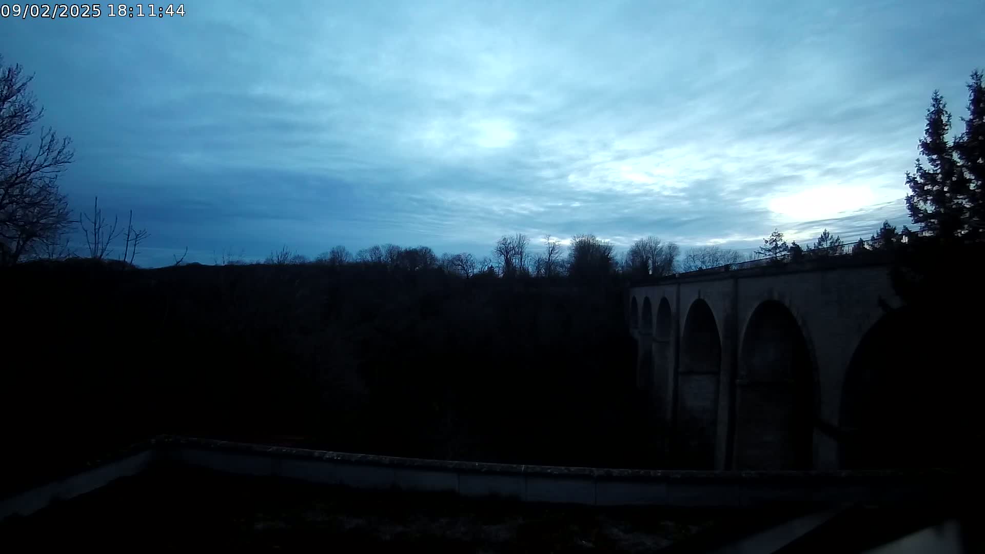 A stone arched bridge is silhouetted against a twilight sky with a mostly cloudy, blue-grey hue.