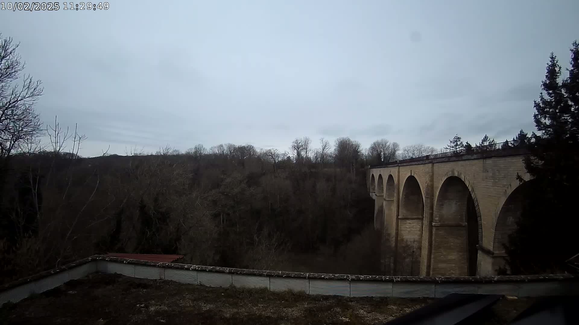 A stone viaduct with multiple arches is visible beyond a line of leafless trees under an overcast sky.