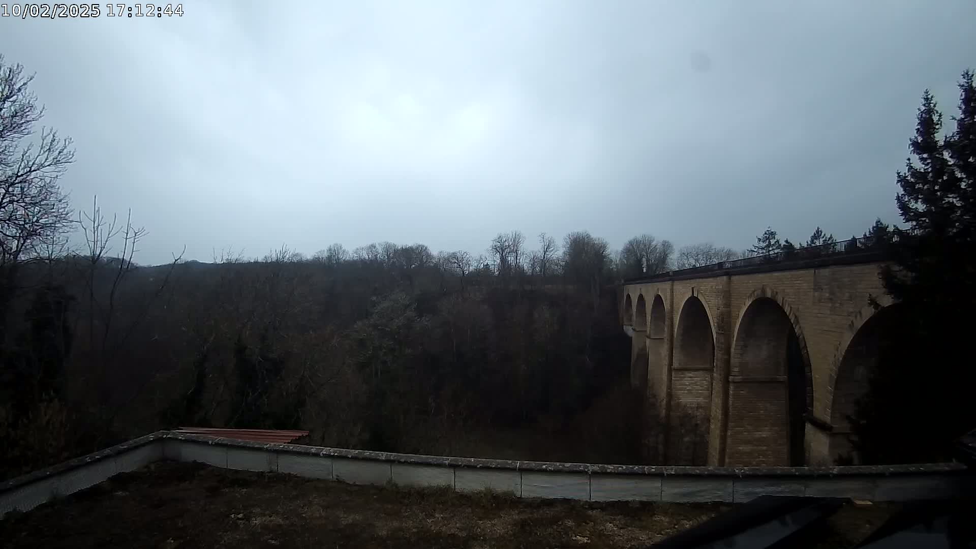 A stone viaduct with multiple arches is visible beyond a low wall and a dark, leafless forest under an overcast sky.