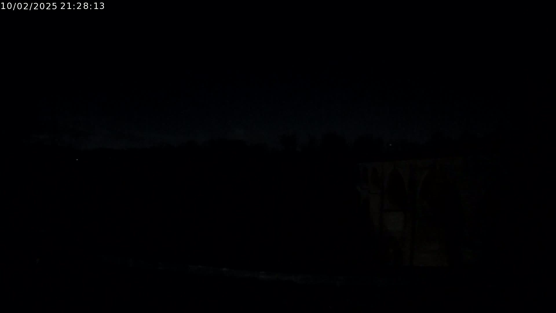 A dark nighttime scene shows a dimly lit structure on the right, with a dark, possibly wooded area to the left under a mostly clear sky.