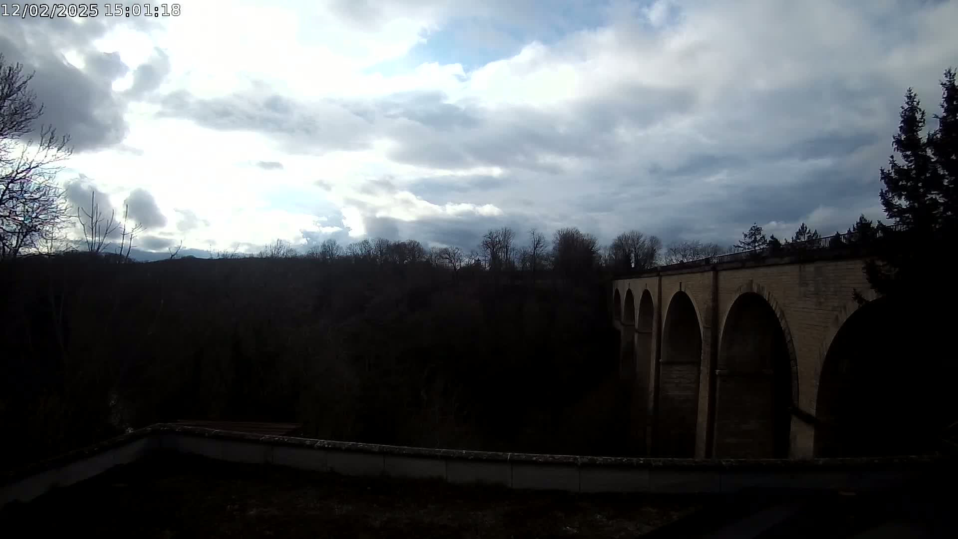 A stone bridge with multiple arches spans a dark valley under a partly cloudy sky.