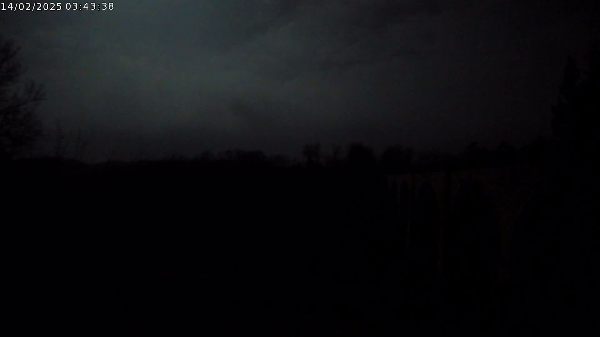 A dark, nighttime scene shows a silhouetted treeline and a structure that appears to be a bridge or viaduct under a mostly cloudy sky.