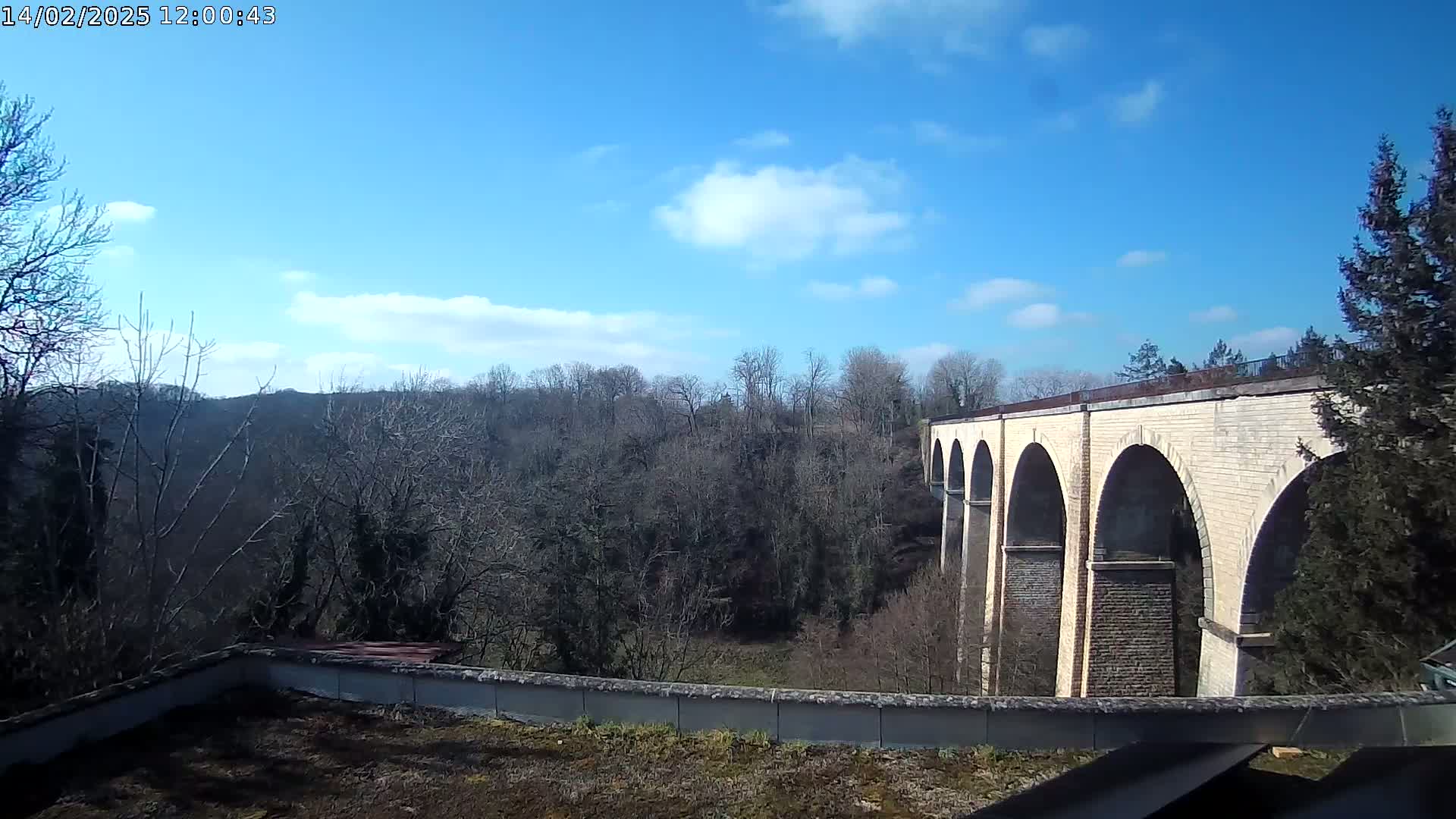 A stone viaduct with multiple arches stretches across a valley of leafless trees under a mostly sunny sky with a few scattered clouds.