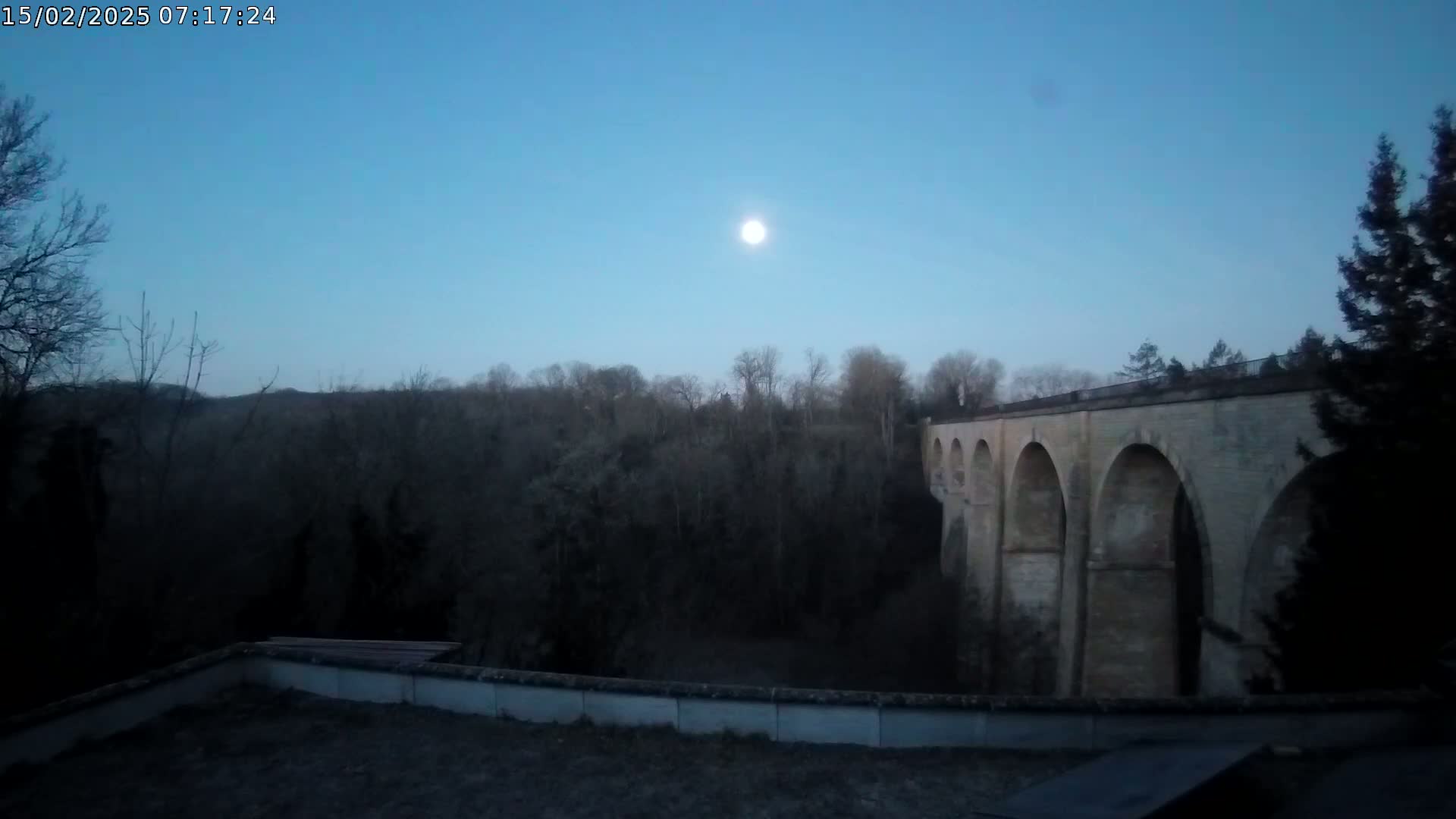 A stone viaduct is visible under a bright sun in a clear blue twilight sky above a dark, leafless forest.