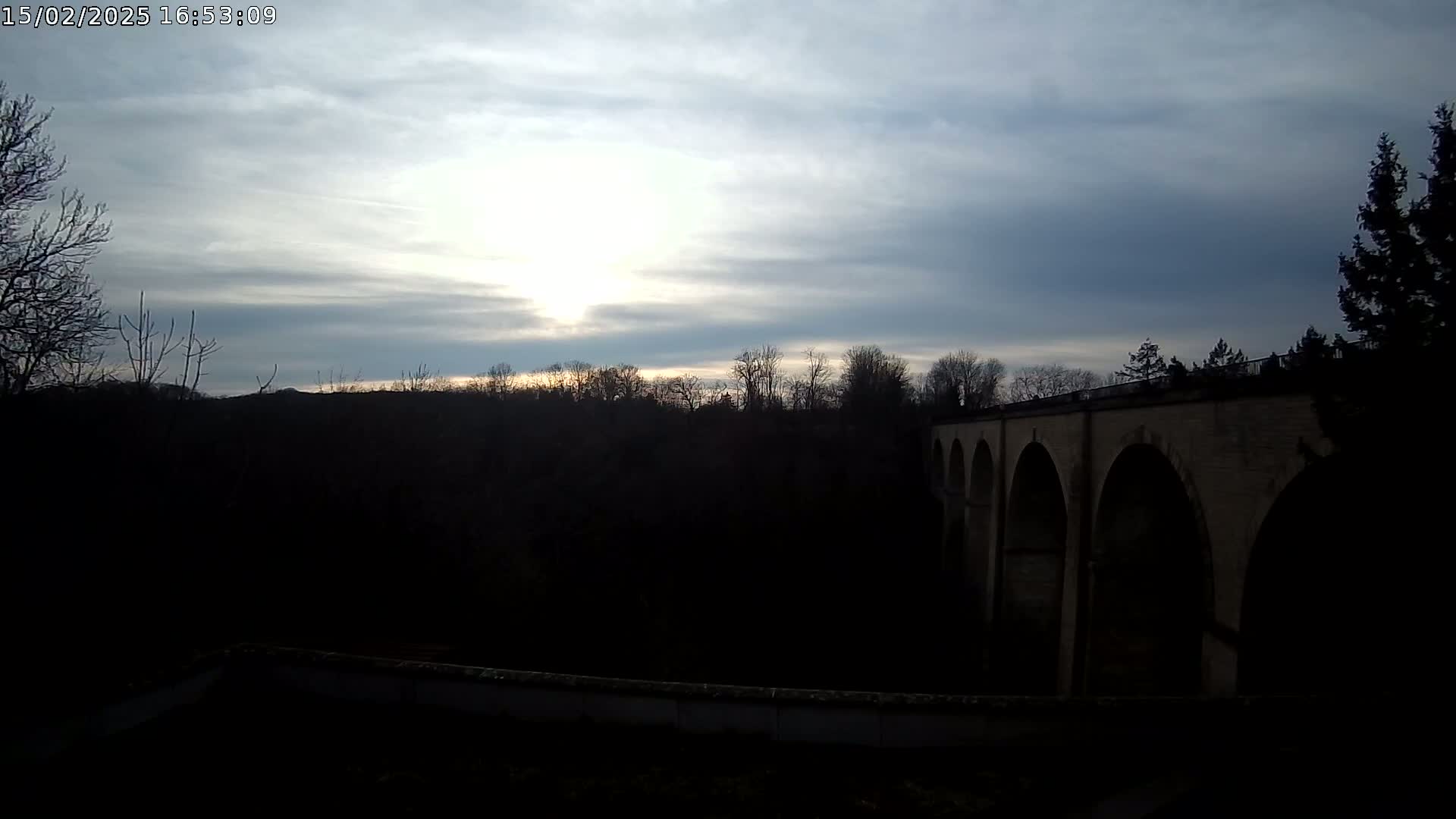 A stone arched bridge is silhouetted against a partly cloudy sunset sky over a dark, tree-lined landscape.
