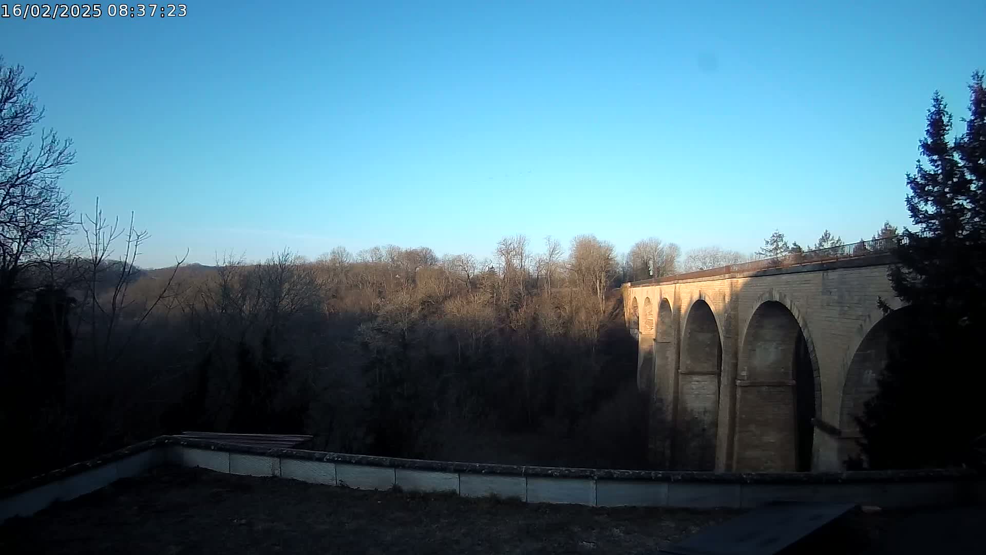 A stone viaduct with multiple arches is visible against a clear blue sky, overlooking a winter woodland.