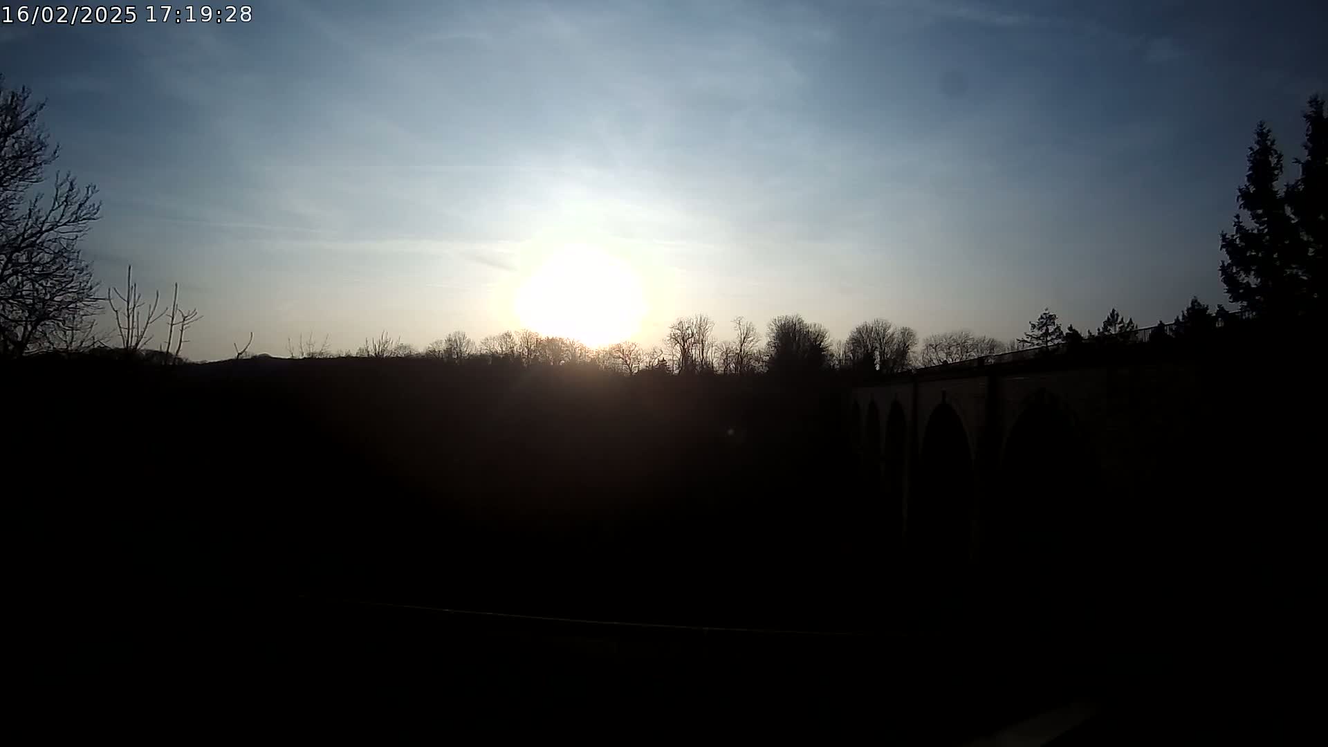 A bright sun sets behind a silhouetted line of trees and a stone bridge under a mostly clear sky.