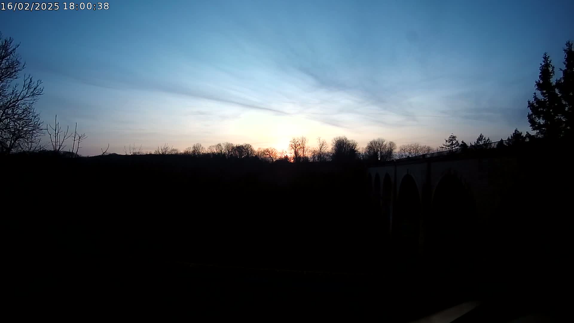 A setting sun casts a warm glow on a silhouetted landscape featuring a stone bridge and leafless trees under a clear, twilight sky.