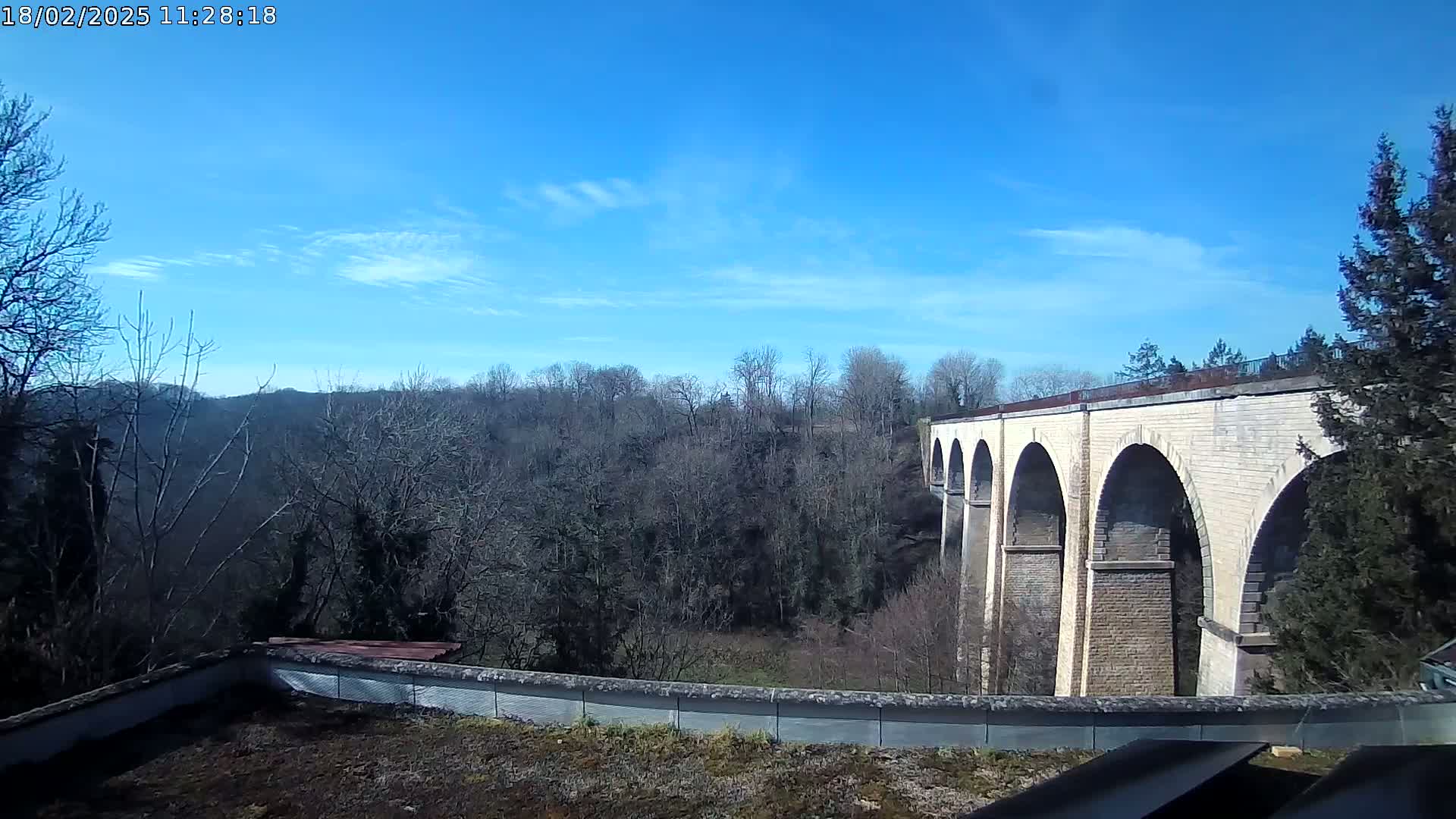 A stone viaduct with multiple arches is visible in the distance, beyond a line of leafless trees under a mostly clear blue sky.