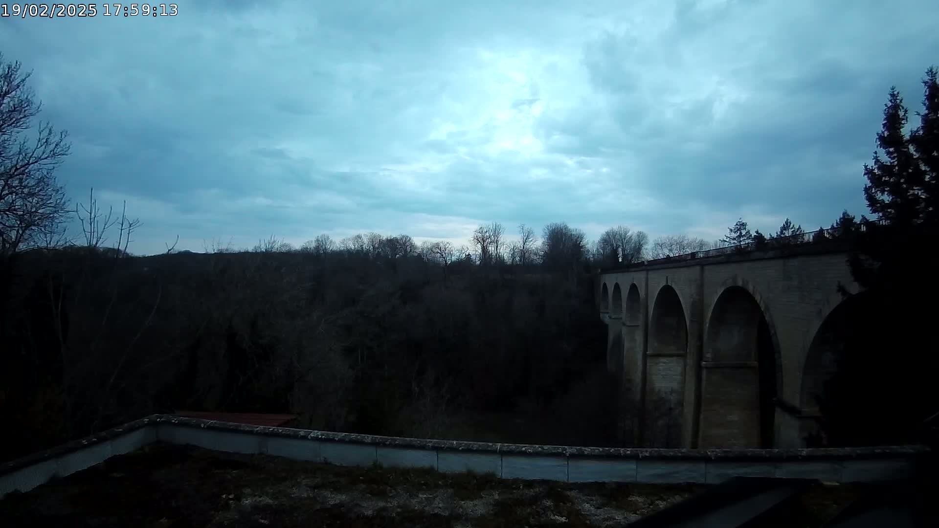 A stone arched bridge is seen beyond a low wall, under a cloudy, dusky sky, with dark trees lining the horizon.