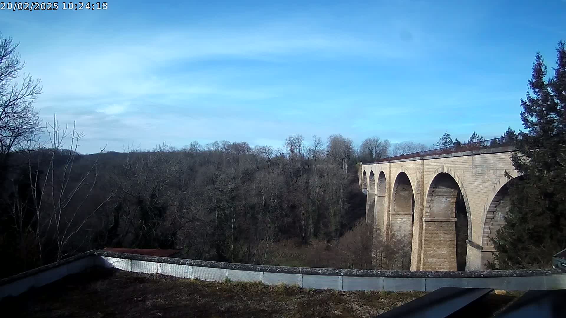A stone viaduct with multiple arches spans a wooded valley under a mostly clear blue sky.
