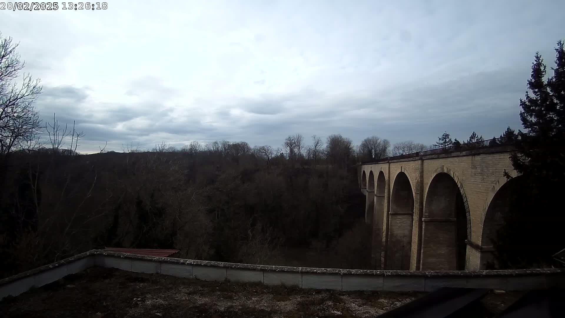 A stone viaduct with multiple arches is seen from a rooftop overlooking a dark, wooded valley under a cloudy sky.