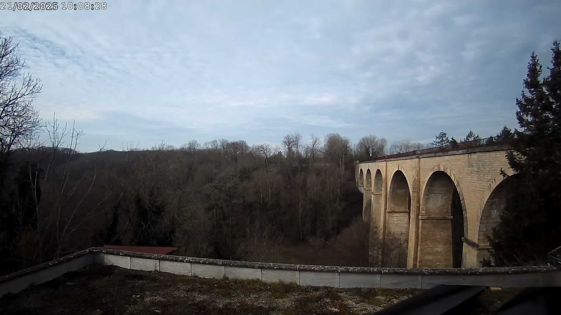 A stone viaduct with multiple arches stretches across a valley of leafless trees under a mostly cloudy sky.