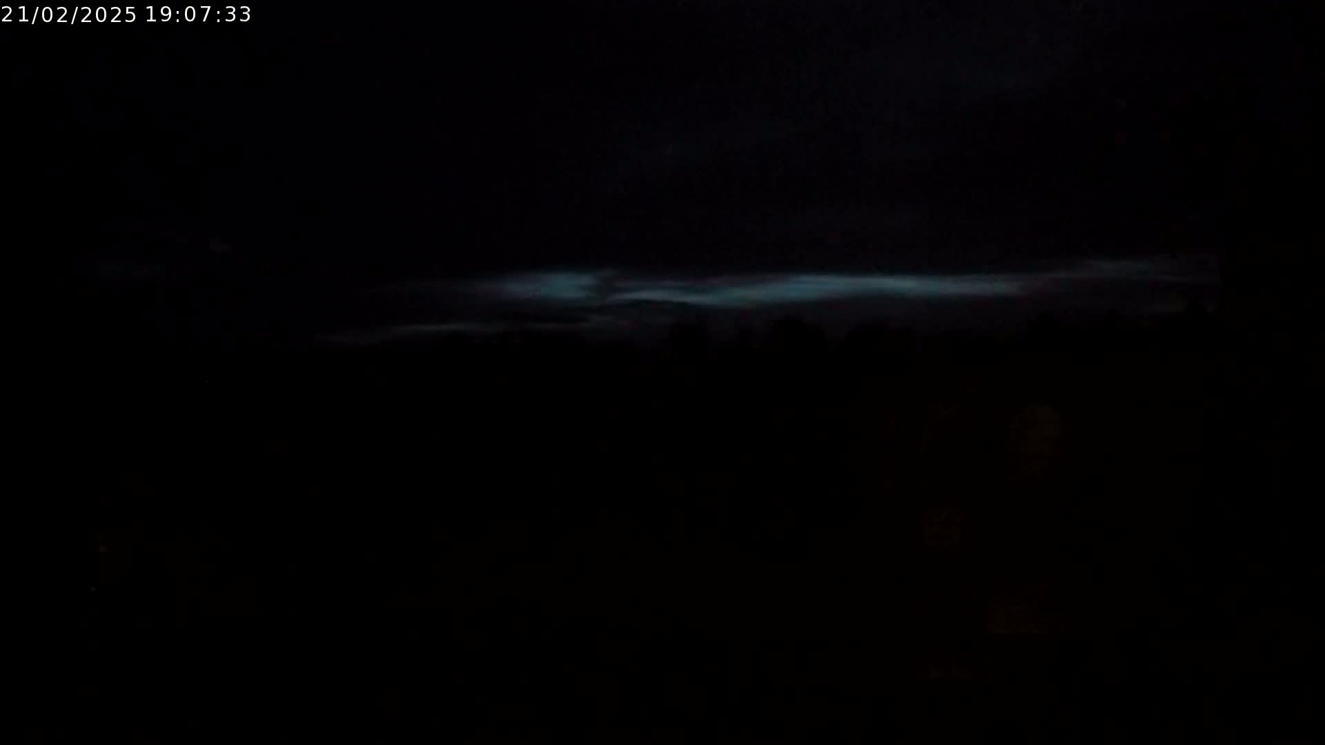 A faintly luminous, horizontal band of clouds is visible in a dark night sky above a silhouetted, treeline.