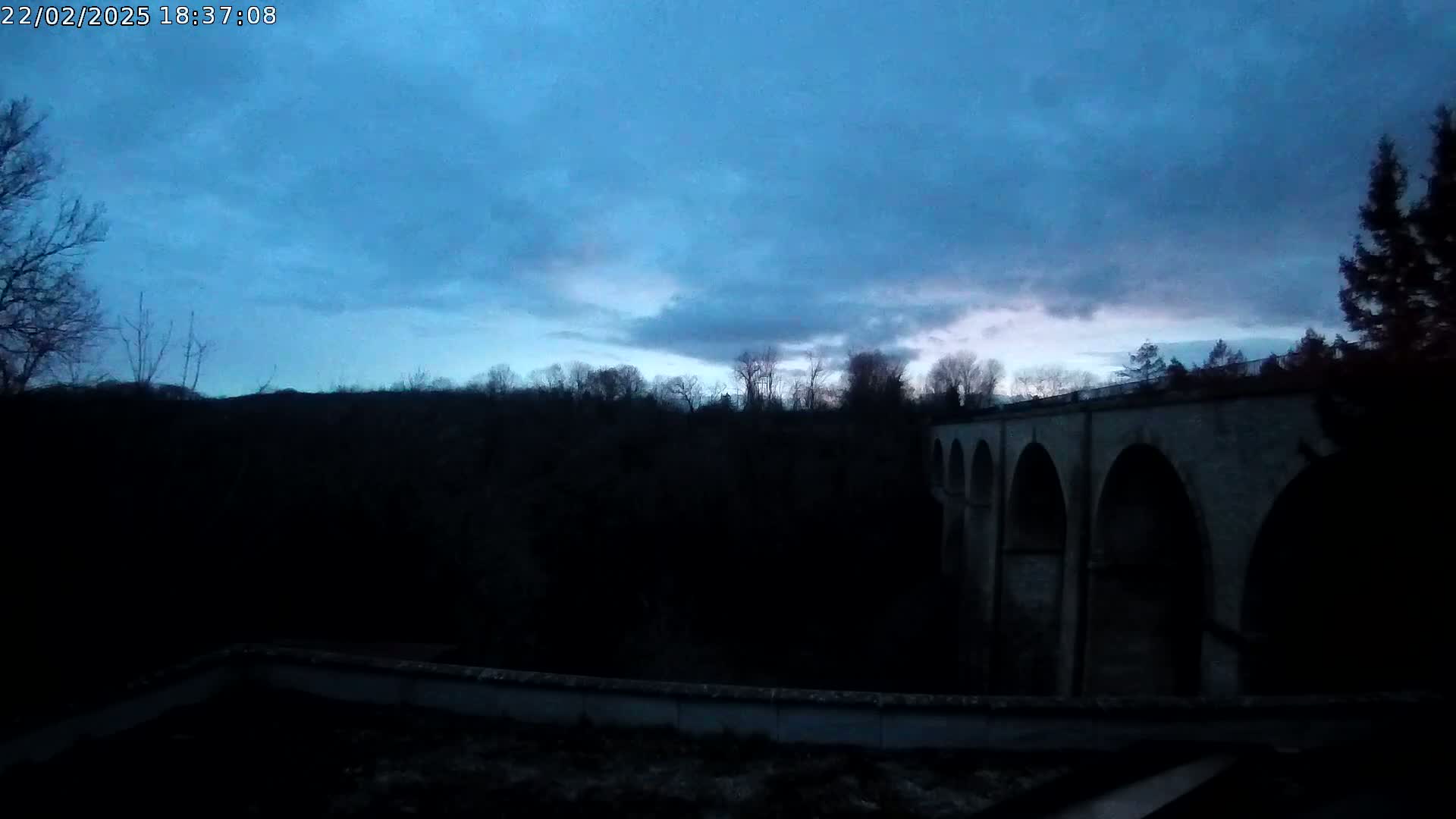 A stone arch bridge is seen at dusk under a mostly cloudy, blue sky.