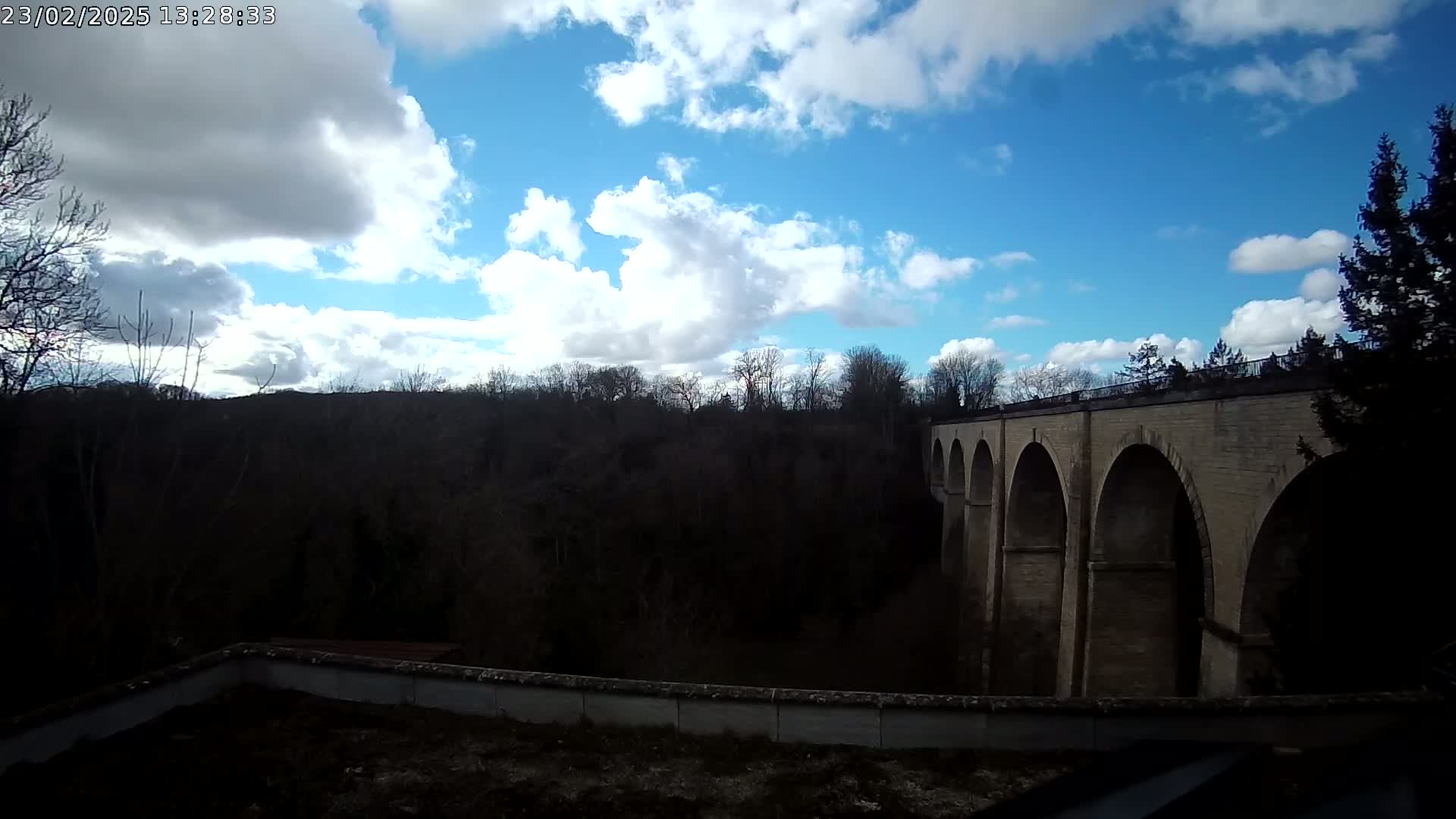 A stone arched bridge stretches across a valley under a partly cloudy blue sky.