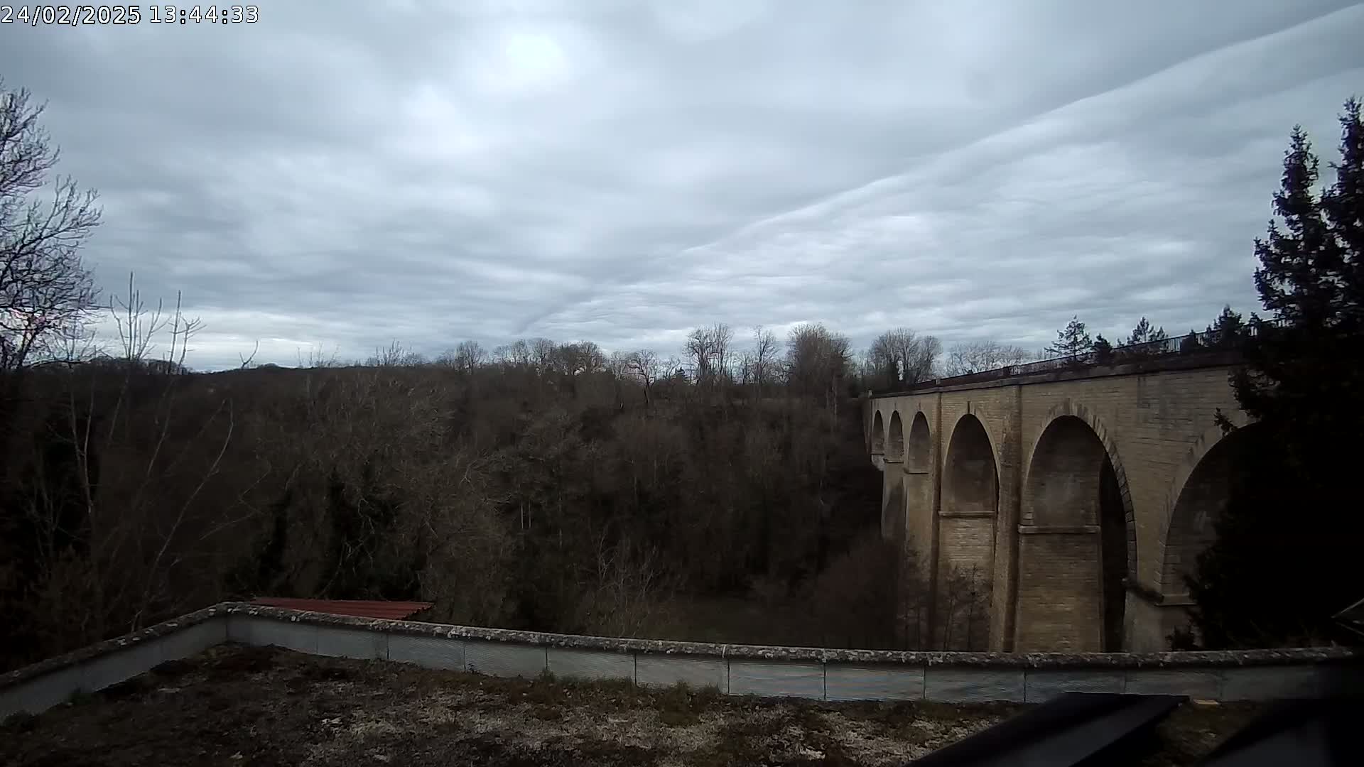 A stone arched viaduct is seen extending from a wooded area under a cloudy sky.