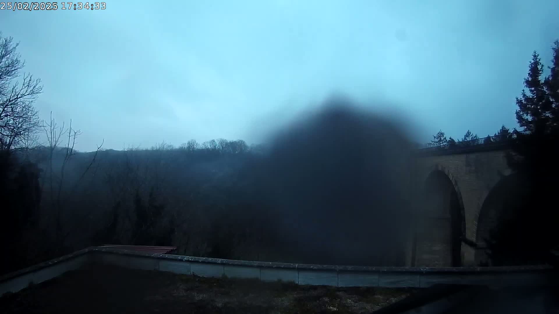 A foggy view shows a stone arch bridge behind a low wall, with trees and a hill shrouded in mist in the background.