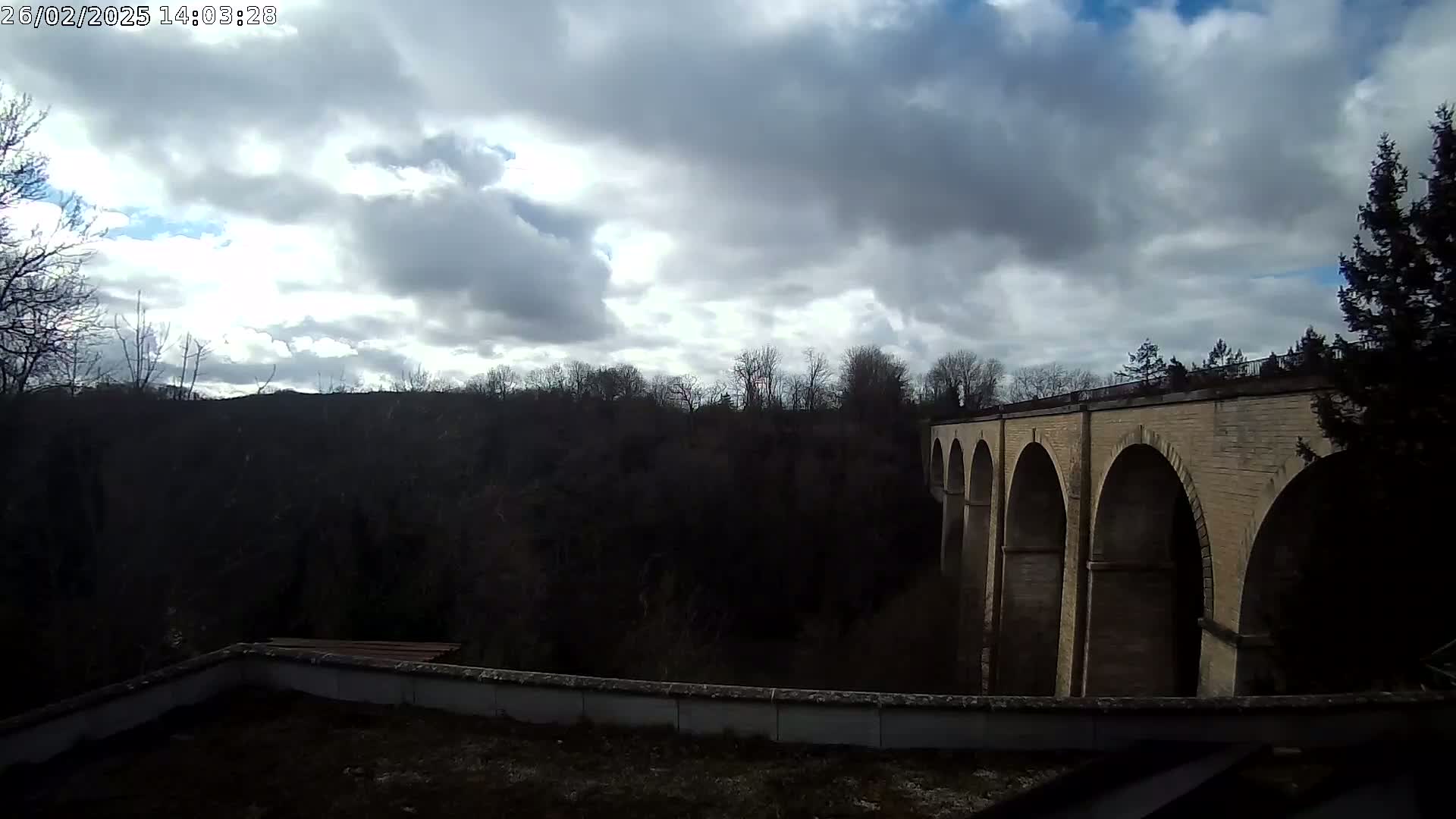 A stone arched viaduct stretches across a wooded valley under a partly cloudy sky.