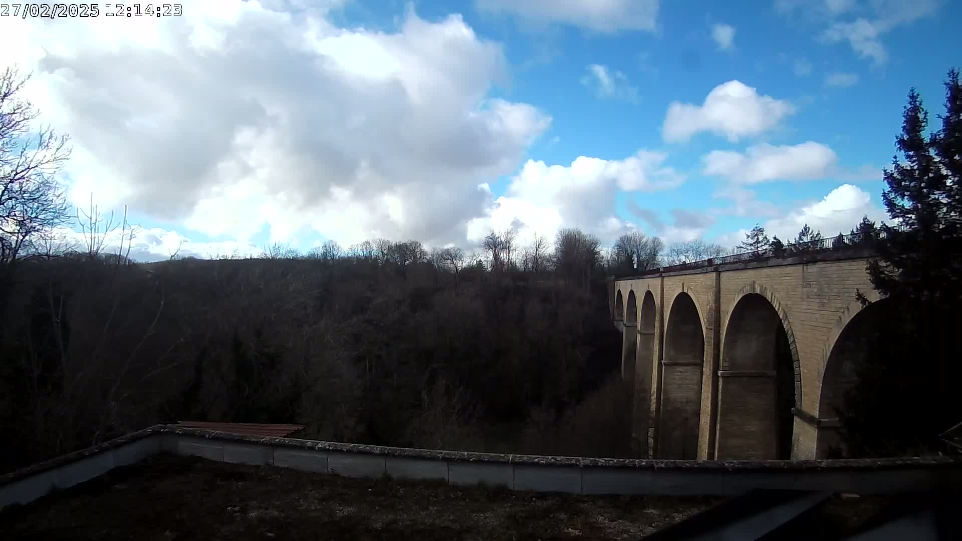 A stone viaduct with multiple arches stretches across a valley of leafless trees under a partly cloudy blue sky.