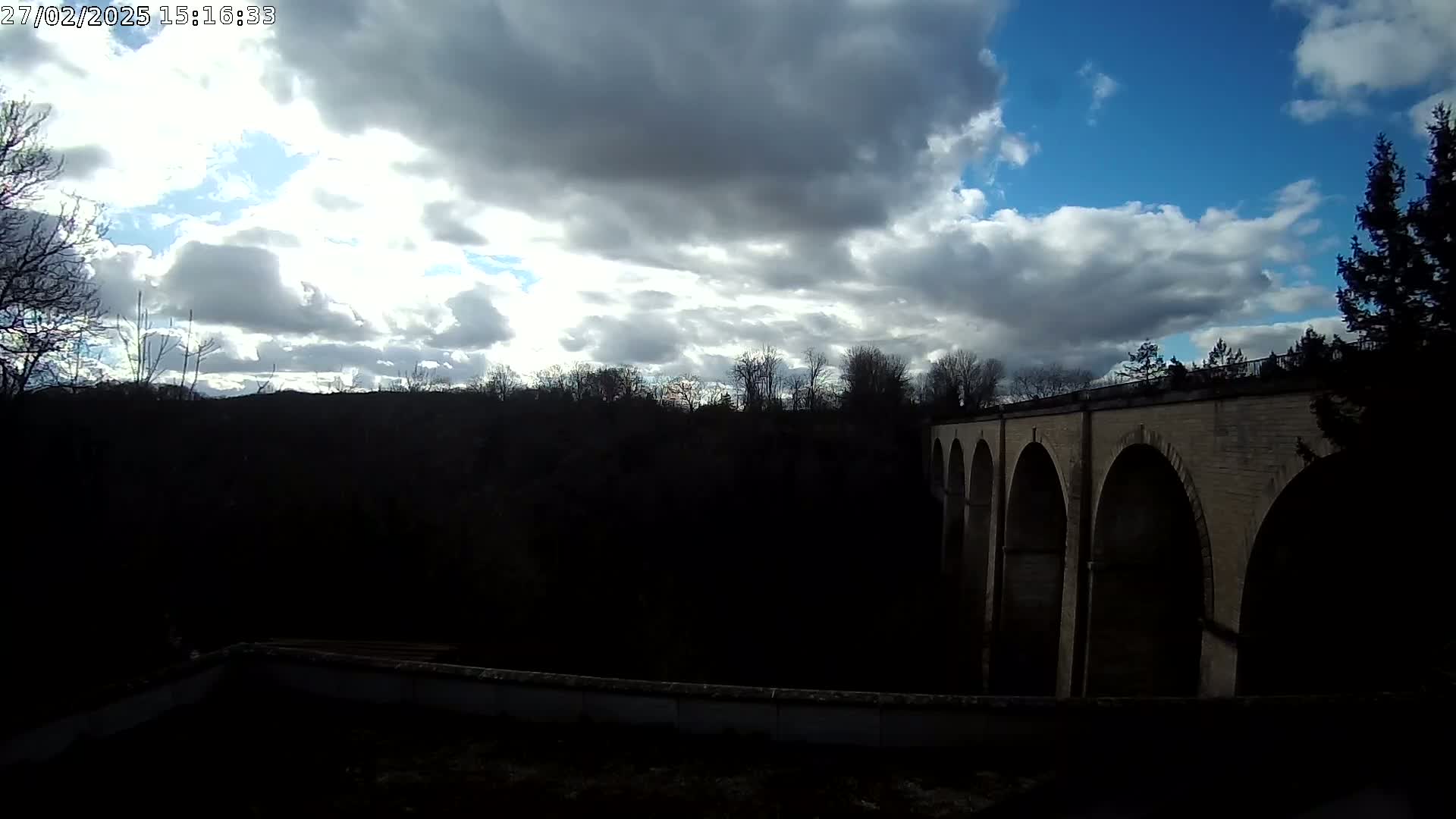 A stone arch bridge stretches across a dark valley under a partly cloudy sky.