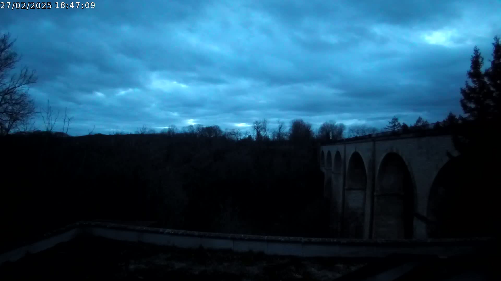 A large stone bridge with multiple arches is visible against a dark blue, overcast sky at dusk.