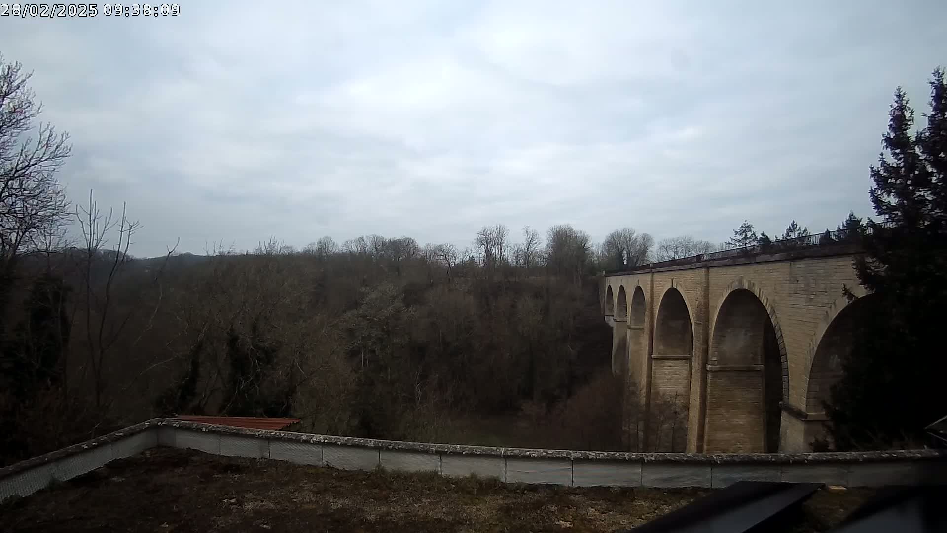 A stone viaduct with multiple arches is visible beyond a low wall and a wooded area under a cloudy sky.