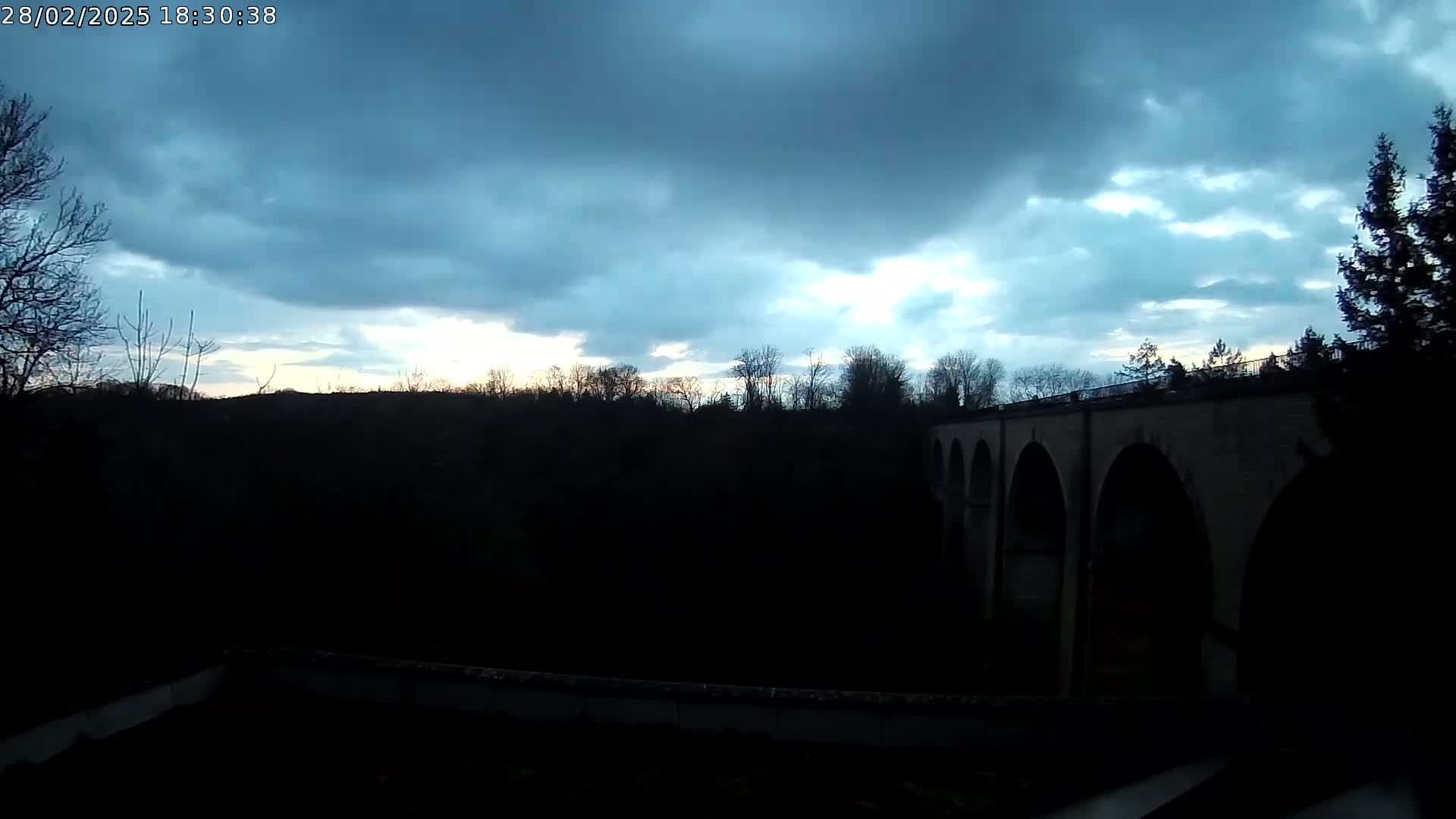 A stone arched bridge is silhouetted against a twilight sky with partly cloudy conditions.